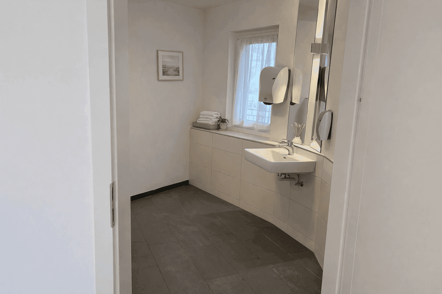 Geräumiger Toilettenbereich mit Fliesenboden, Waschbecken und Spendersystemen an der Wand.