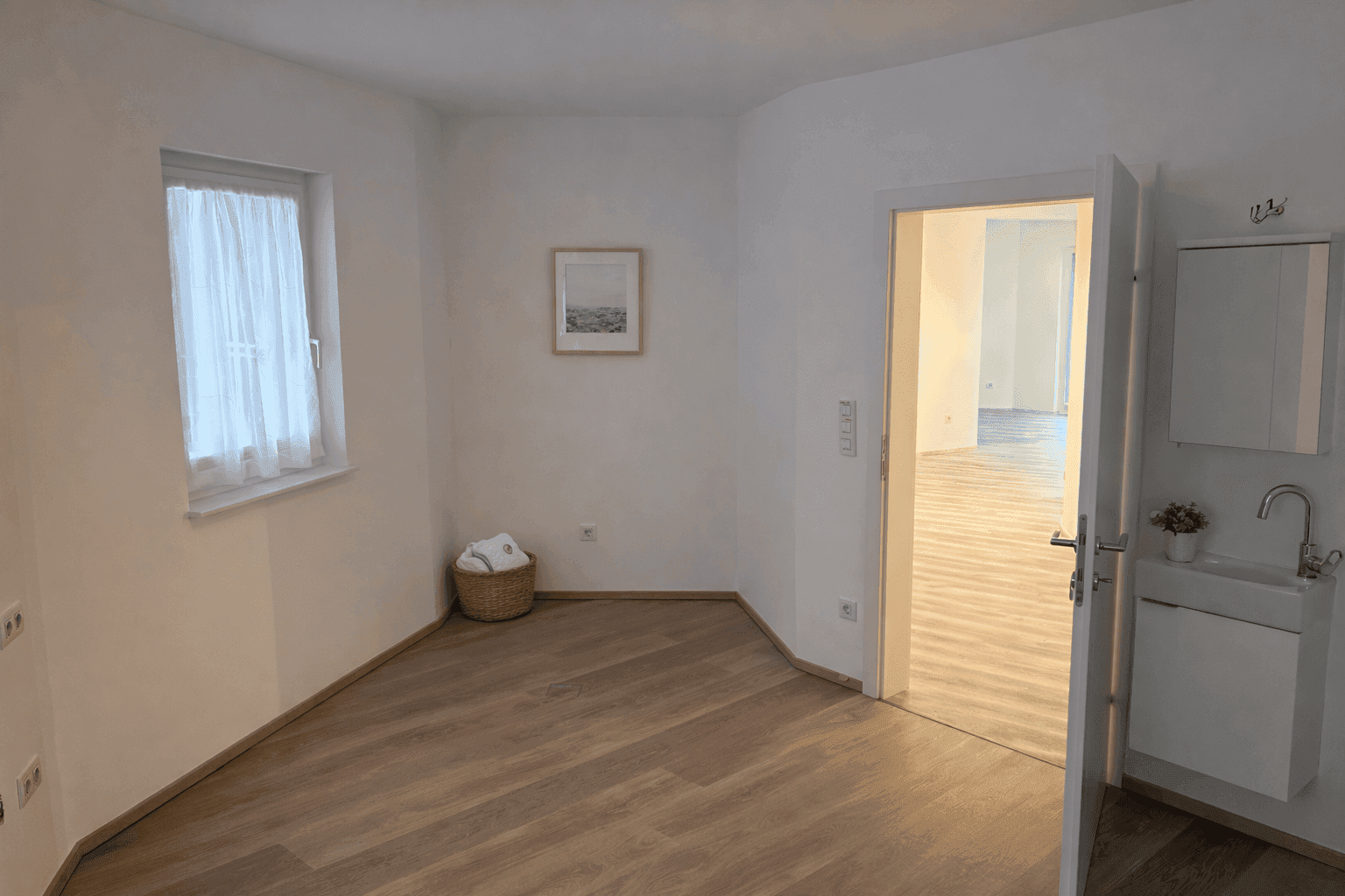 Helles Zimmer mit Laminatboden, einem Fenster mit Vorhang und Zugang zu einem weiteren Raum.