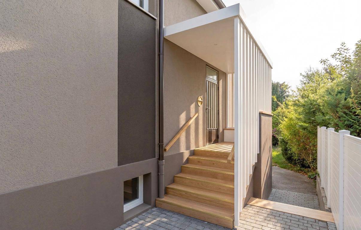 Moderner Hauseingang mit Holztreppe und Überdachung, umgeben von Grünflächen.