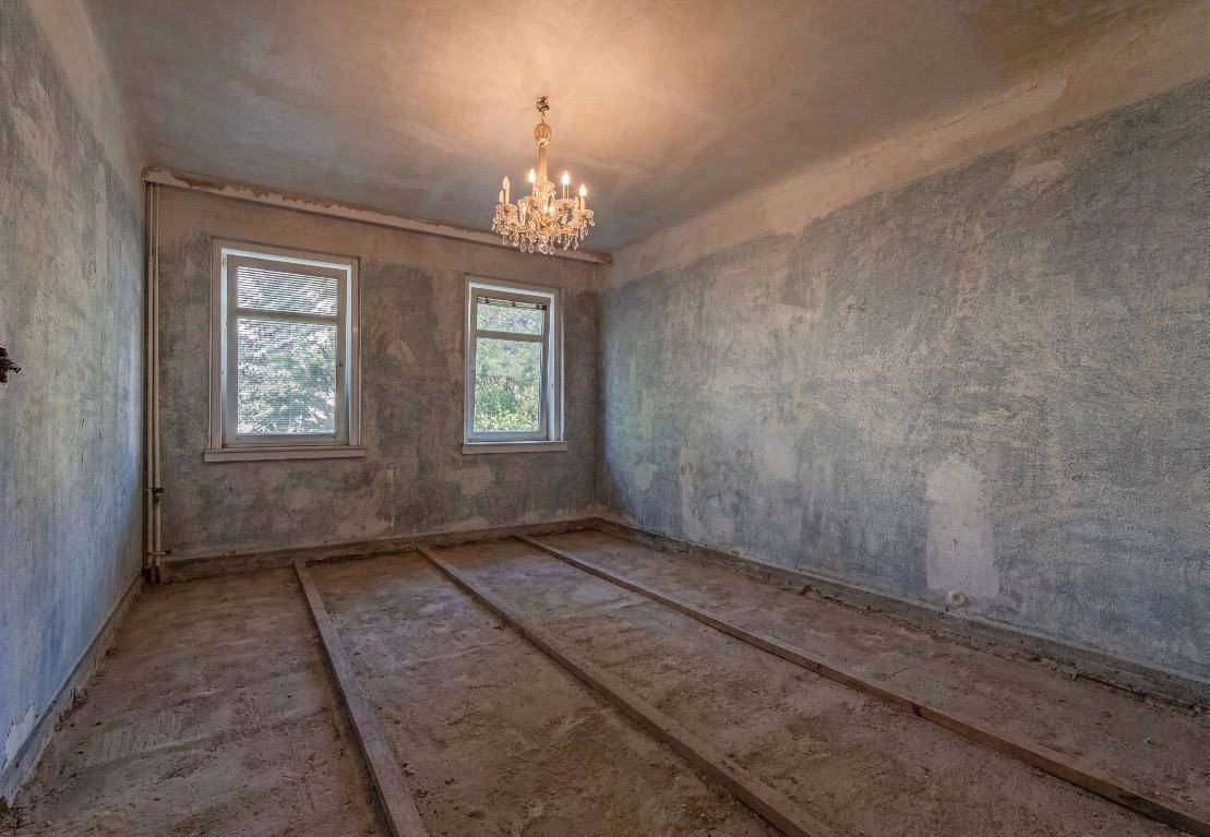 Unausgebautes Zimmer mit zwei Fenstern und einem klassischen Kronleuchter, bereit für die Gestaltung.