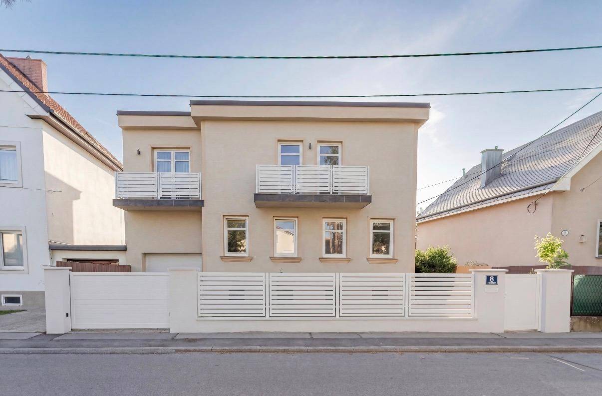 Renovierte Hausfassade mit modernen Balkonen und weißem Zaun zur Straße hin.