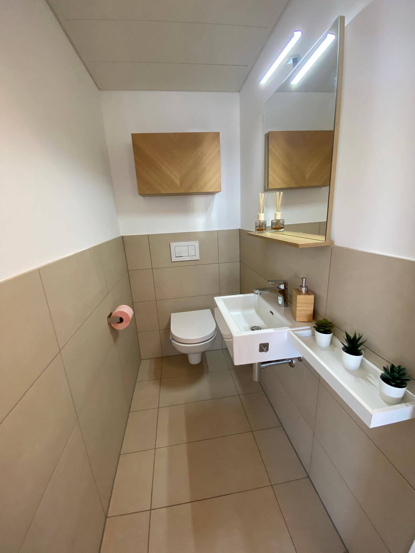 Modernes Gäste-WC mit Wand-WC, kleinem Waschbecken und beigen Fliesen an den Wänden.