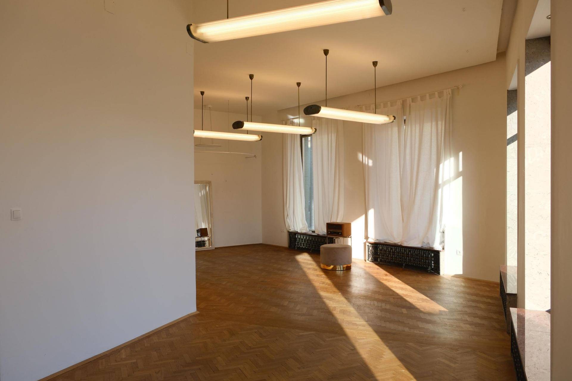 Geräumiger, heller Raum mit Fischgrätparkett, großen Fenstern und modernen Hängelampen, ideal für Ausstellungen.