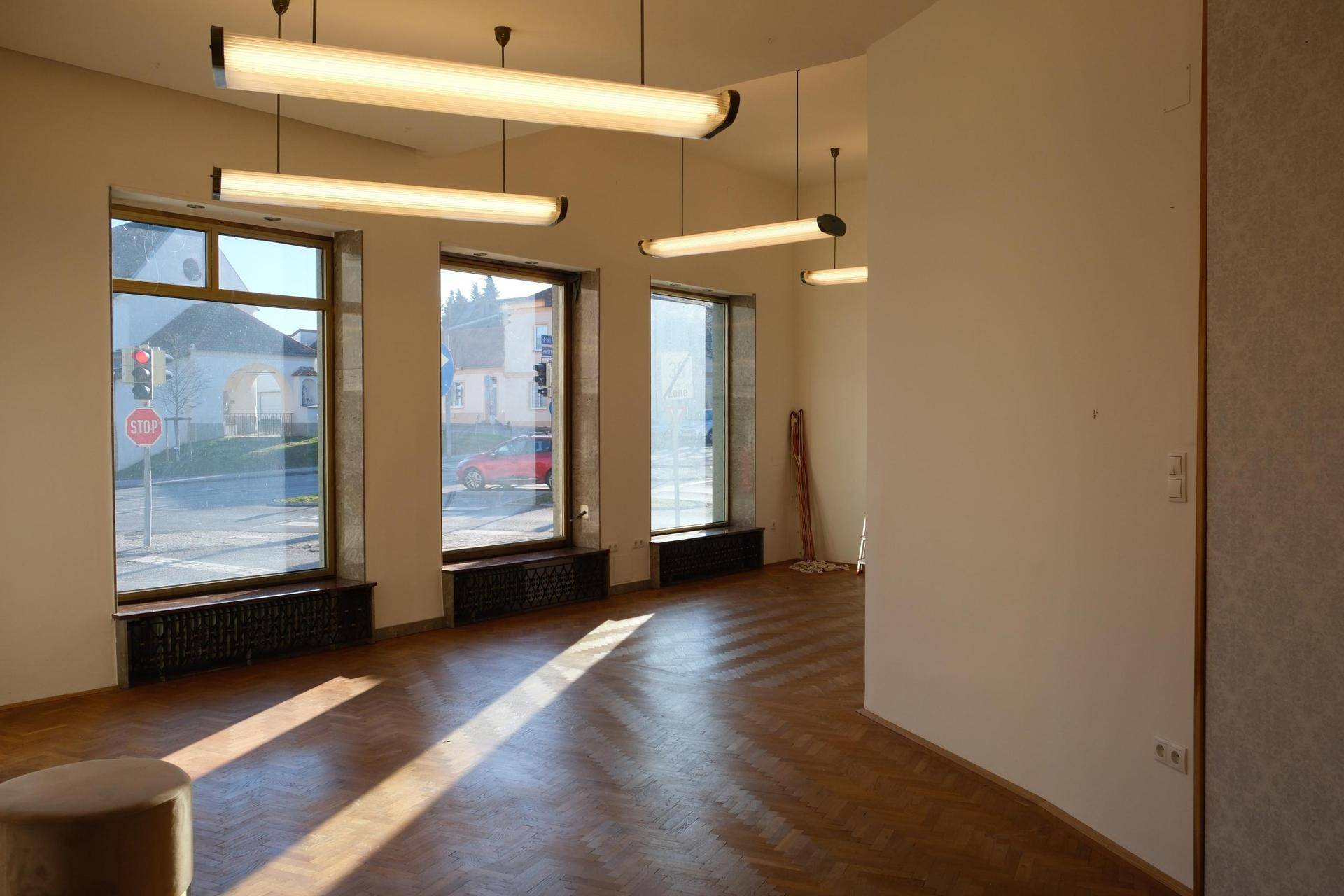 Heller Raum mit Fischgrätparkett und großen Fenstern, die viel Tageslicht hereinlassen und einen Blick auf die Straße bieten.