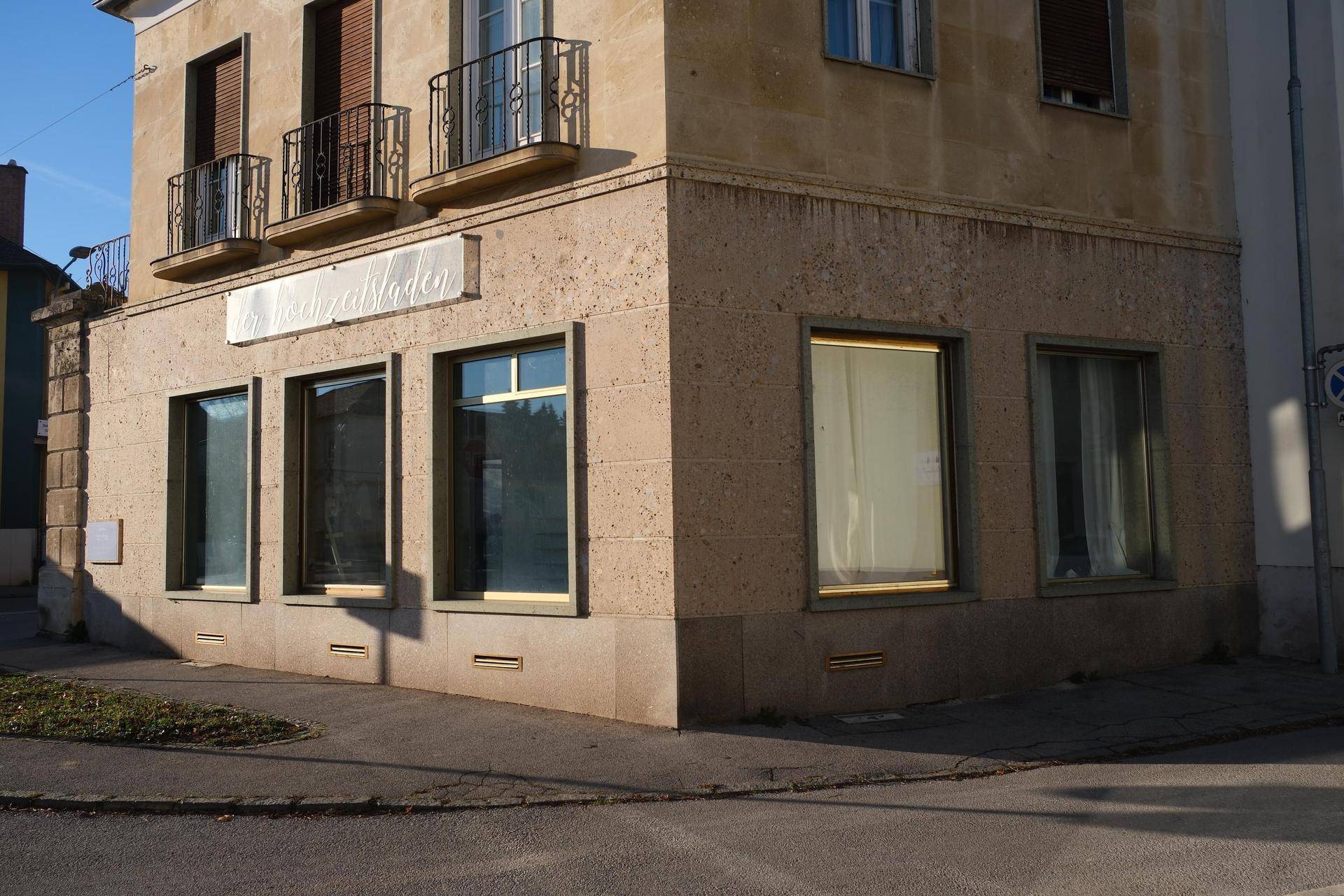 Unterer Bereich eines Eckgebäudes mit großen Schaufenstern und einer robusten Steinfassade, an einer Straßenecke.