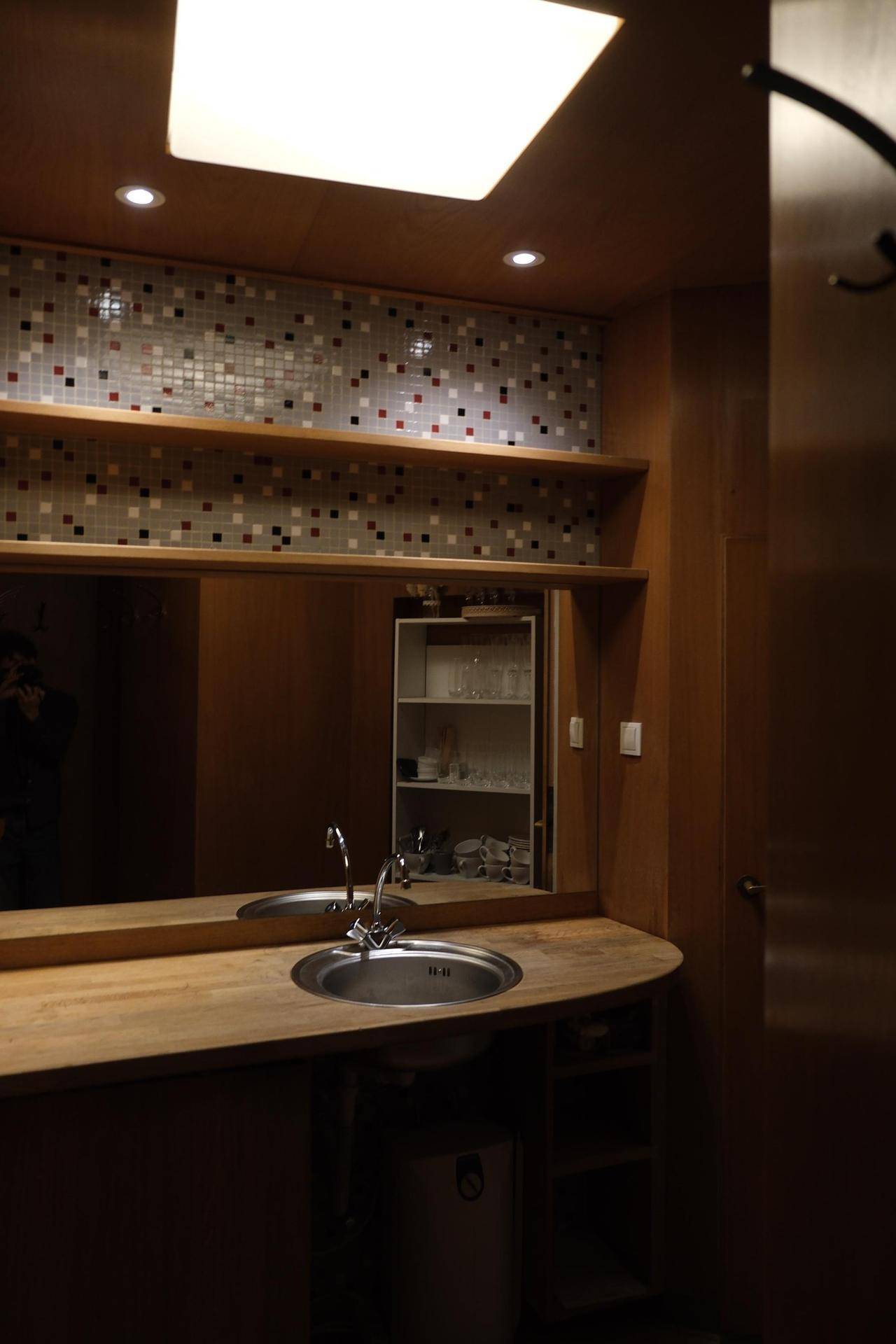 Waschbereich mit Holztheke, rundem Waschbecken und großem Spiegel, beleuchtet von einer Deckenleuchte.