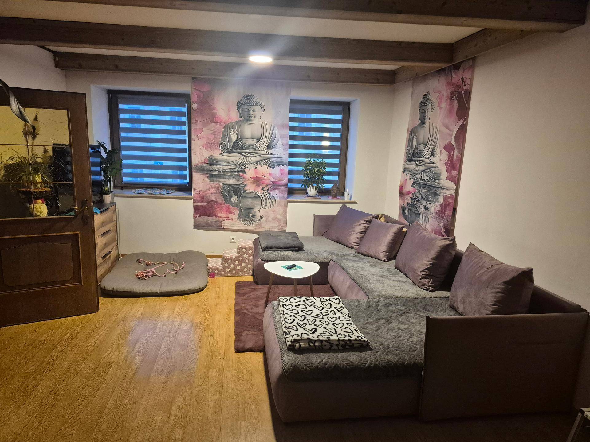 Wohnzimmer mit großem Ecksofa, Holzboden, sichtbaren Deckenbalken und dekorativen Wandbehängen.