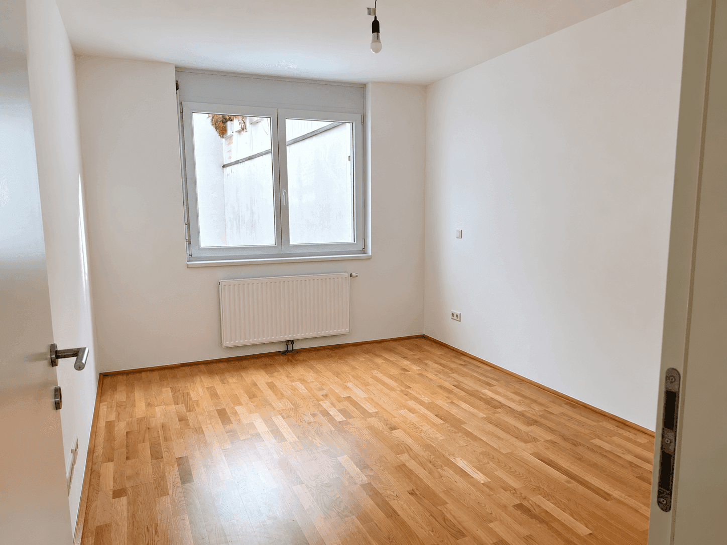 Leeres Schlafzimmer mit Parkettboden, weißen Wänden und Fenster mit Rollladen.