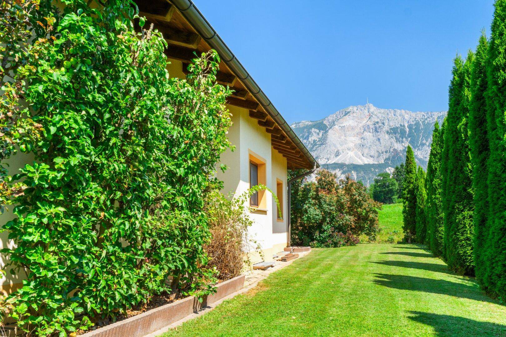 Gepflegter Seitenbereich des Hauses mit Rasenfläche, hoher Hecke und Blick auf die umliegenden Berge.