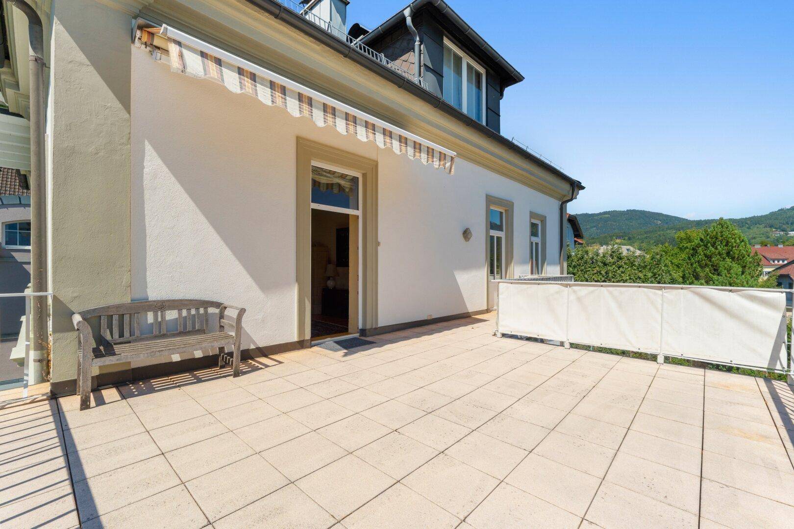 Große, sonnige Terrasse mit Fliesenboden, Sitzbank und Blick auf die Umgebung.