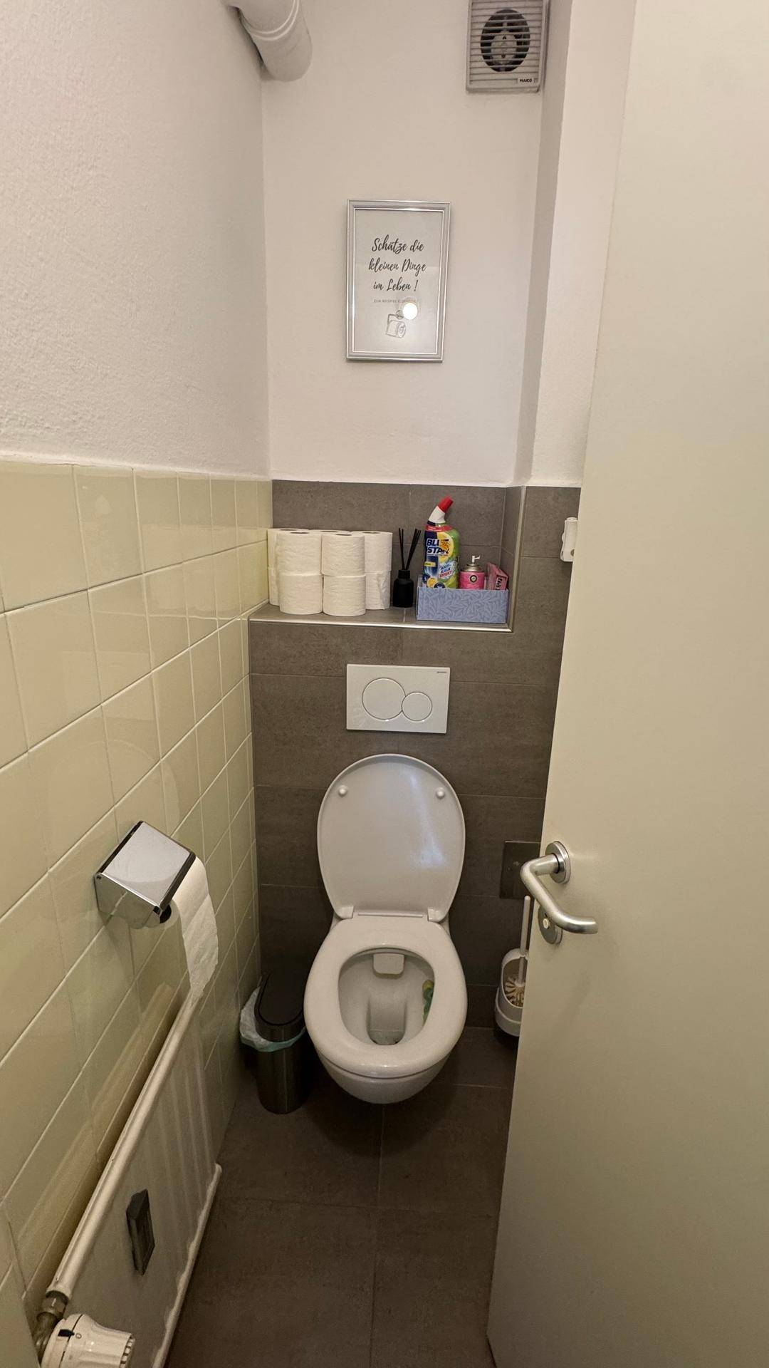 Kleines Gäste-WC mit Wand-WC, halbhohen Fliesen und Abluftventilator.