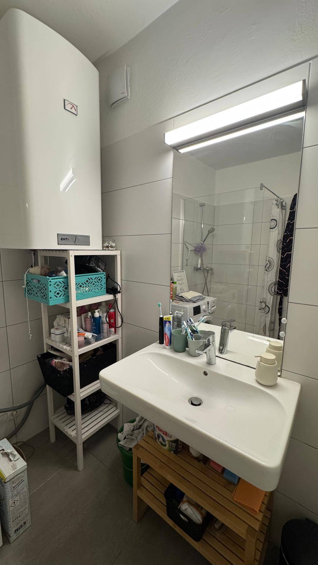 Badezimmer mit Waschbecken, Spiegel und einem Warmwasserboiler über einem Regal.
