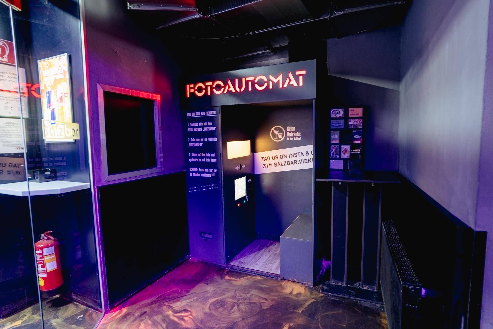 Eingangsbereich mit einem Fotoautomaten und Hinweisschildern, beleuchtet von violettem Neonlicht.