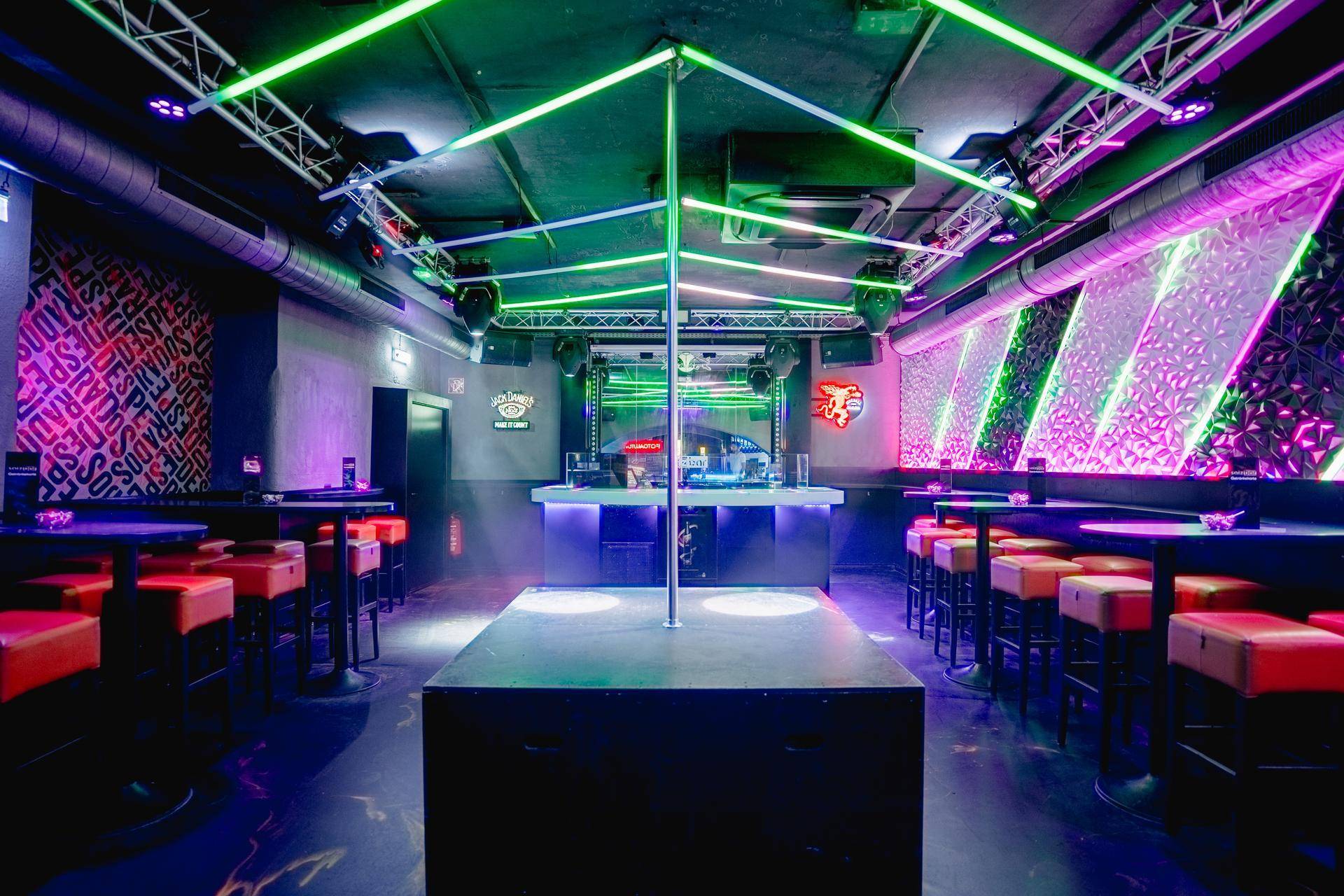 Dynamischer Clubbereich mit Tanzstange, DJ-Pult und Sitzgelegenheiten, beleuchtet von grünen und violetten Neonlichtern und Rauch.