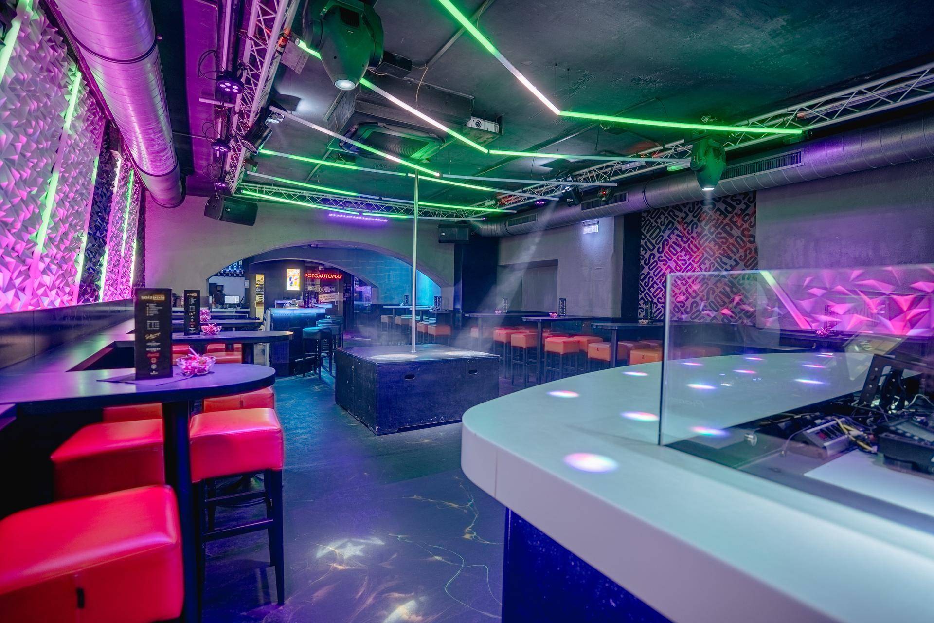 Blick vom DJ-Pult über den Clubbereich mit Tanzstange und Sitzgelegenheiten bis zur beleuchteten Bar im Hintergrund.