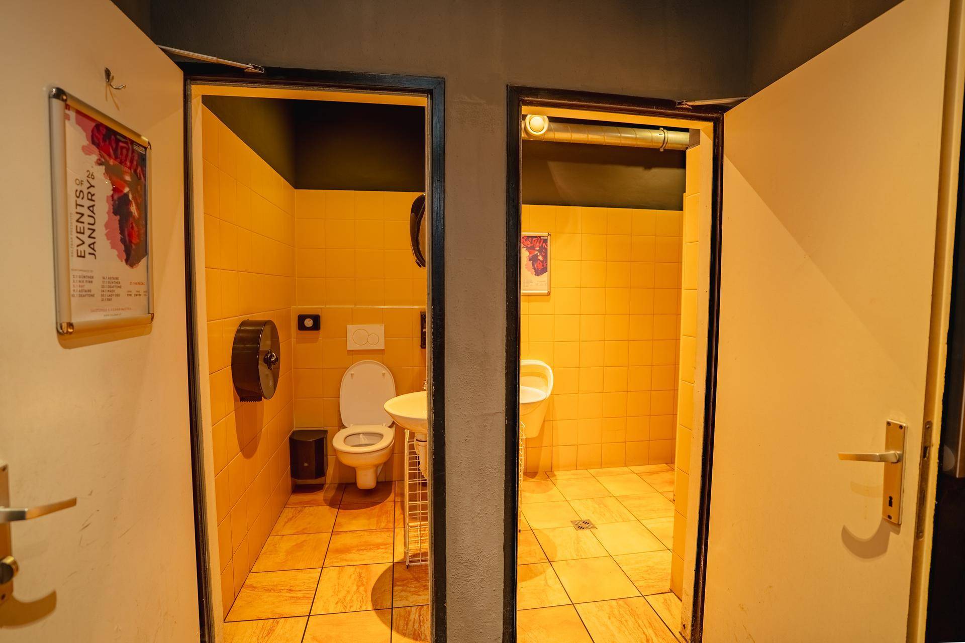 Zwei separate Toilettenräume mit gelben Fliesen, jeweils ausgestattet mit WC und Waschbecken oder Urinal.