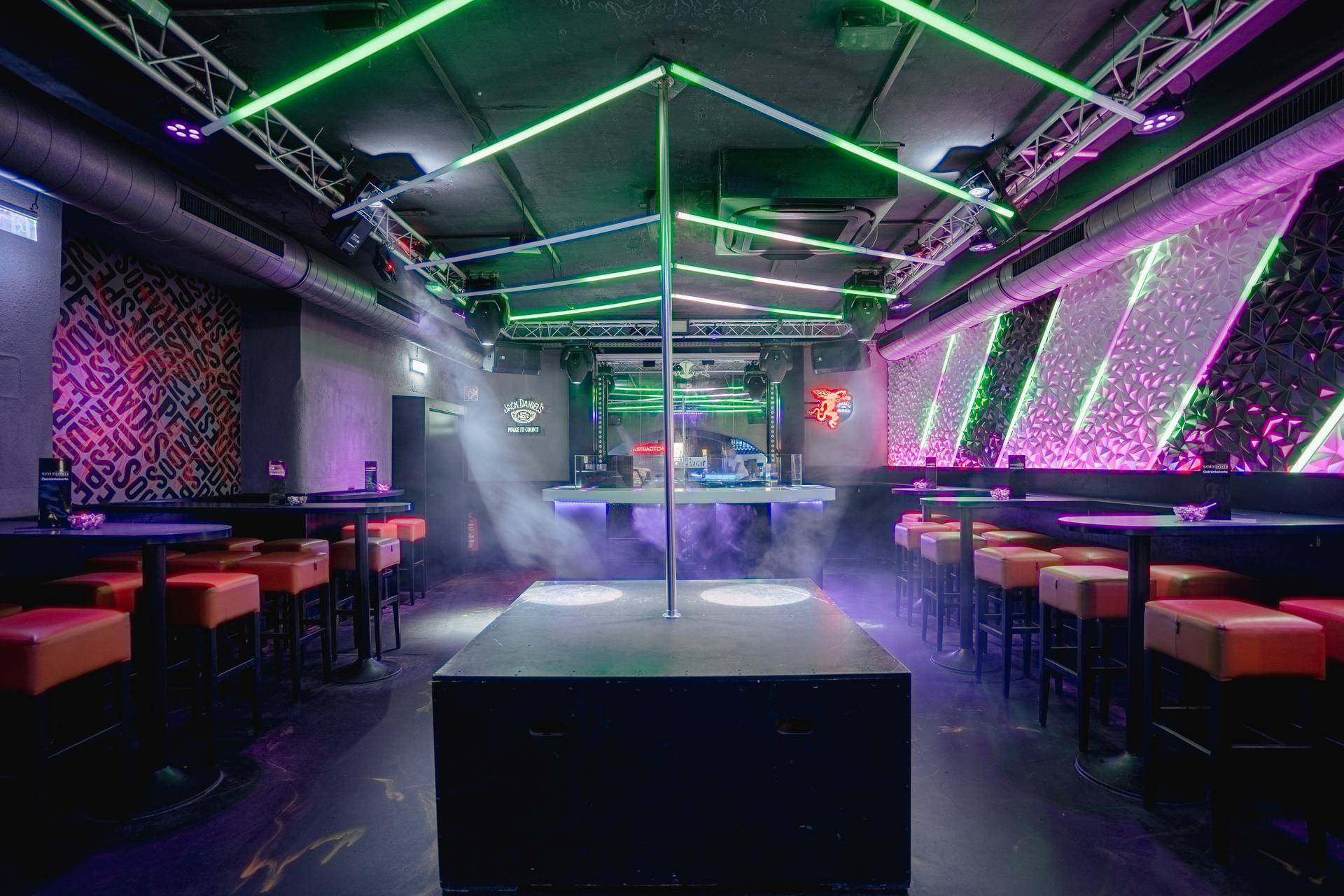 Atmosphärischer Clubraum mit Tanzstange, DJ-Pult und Sitzgelegenheiten, beleuchtet von grünen und violetten Lichtern mit Raucheffekten.