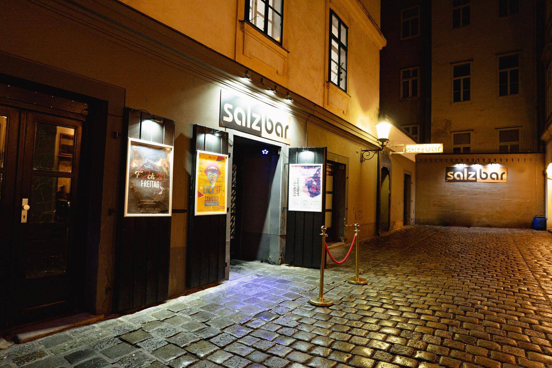 Nachtansicht des Eingangs zur Salzbar mit Kopfsteinpflaster, Laternen und Werbeplakaten an der Fassade.