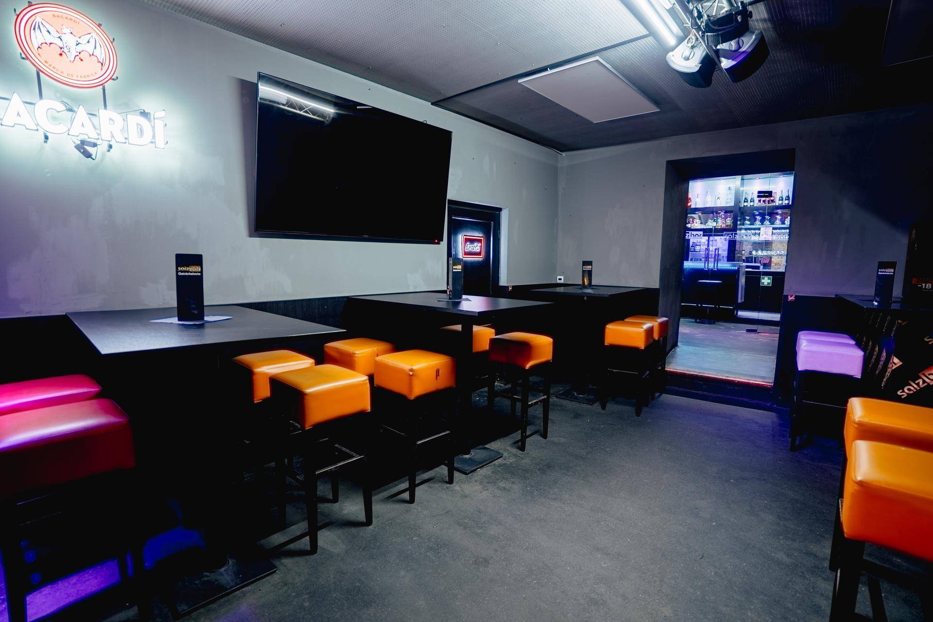 Gemütlicher Lounge-Bereich mit Tischen, orangefarbenen Hockern und einem großen Wandfernseher, ideal für gesellige Abende.