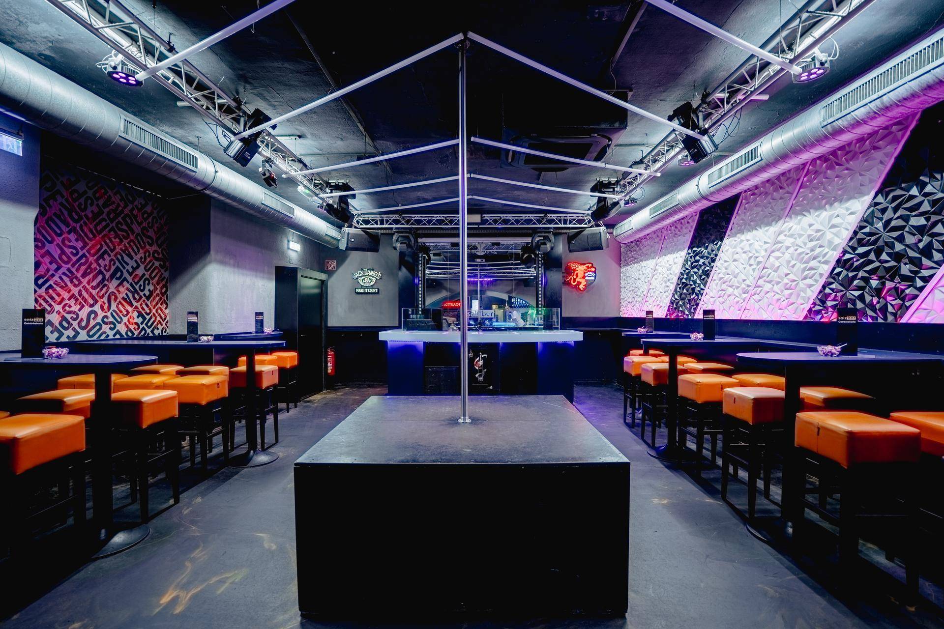 Hauptbereich der Bar mit Tanzstange, DJ-Pult und Sitzgelegenheiten in Schwarz und Orange unter Bühnenbeleuchtung.