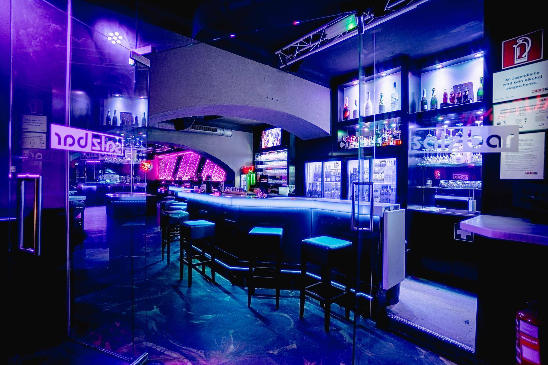 Eingangsbereich der Bar mit beleuchteter Theke, Glaswänden und reflektierenden Böden unter Neonlicht.