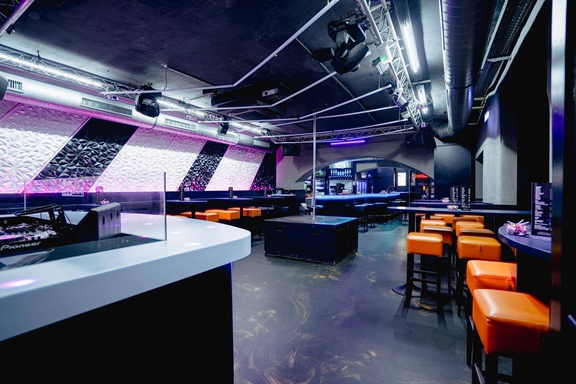 Blick vom DJ-Pult auf den Hauptbereich des Clubs mit Tanzstange und Sitzgelegenheiten, umgeben von modernen Wanddesigns.