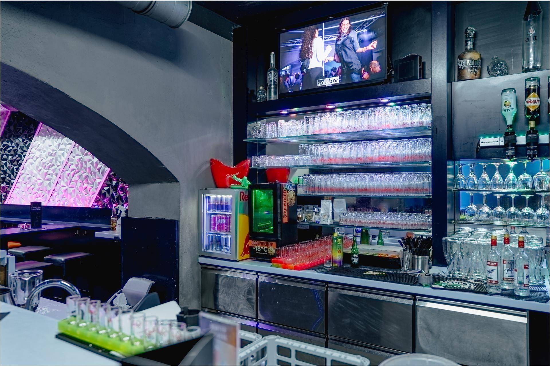 Detailansicht der Bar mit gefüllten Gläserregalen, Kühlschränken und einem Fernseher über der Theke.