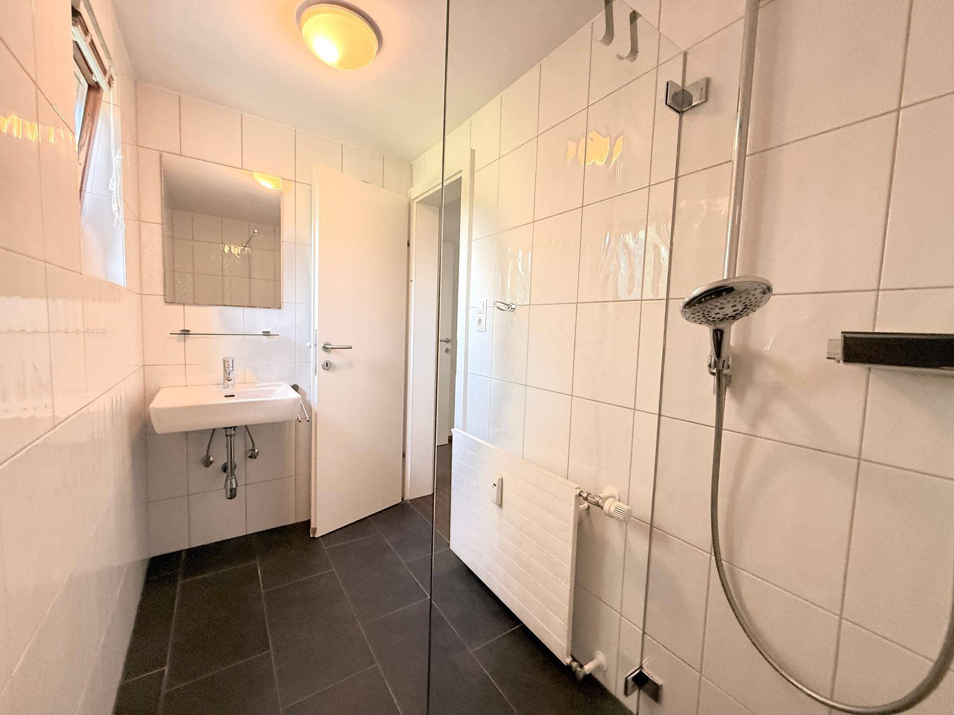 Modernes Badezimmer mit weißen Wandfliesen, dunklem Boden und einer begehbaren Dusche mit Glasabtrennung.