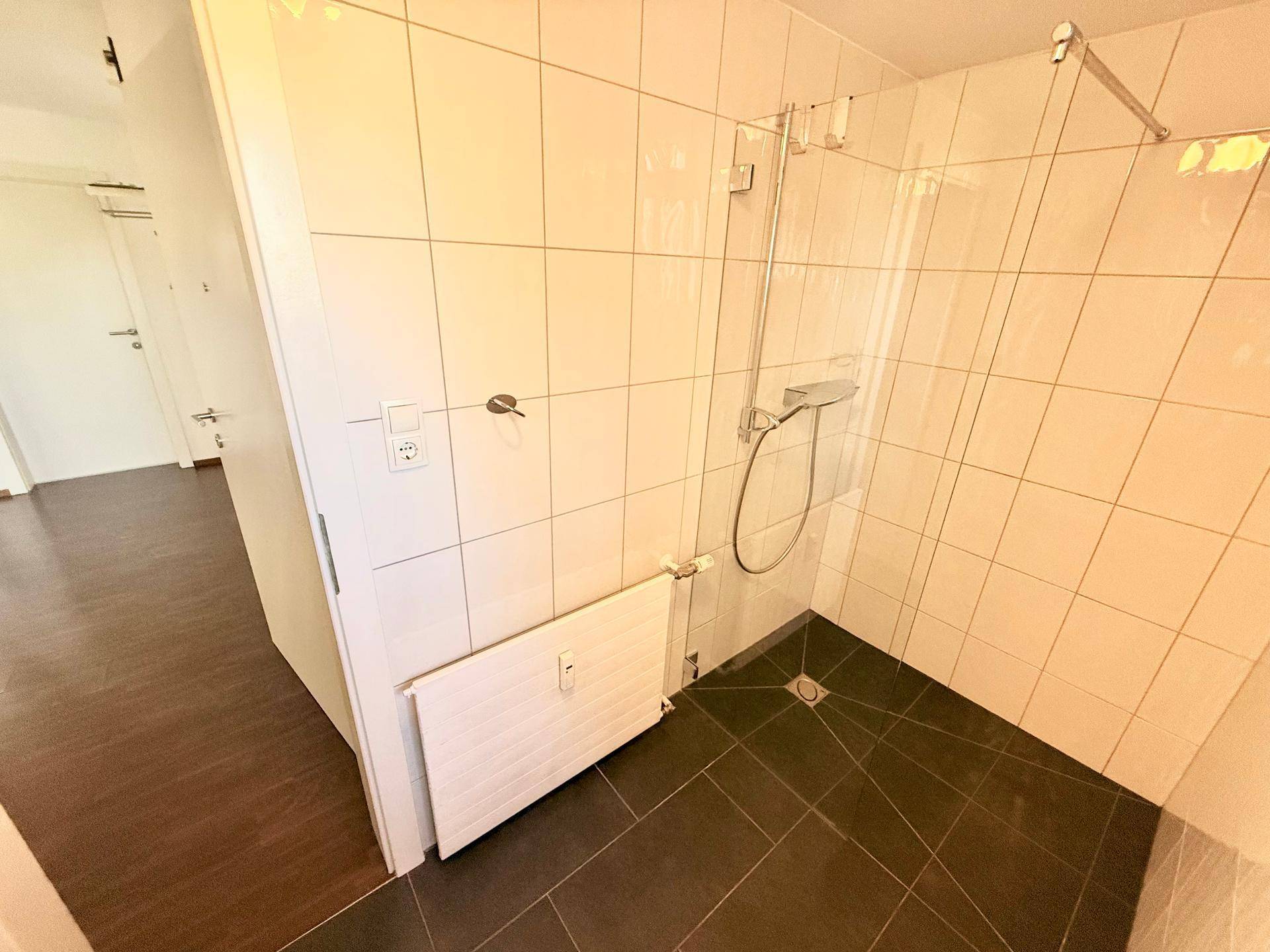 Geräumige Dusche mit Glasabtrennung und dunklem Fliesenboden im hell gefliesten Badezimmer.