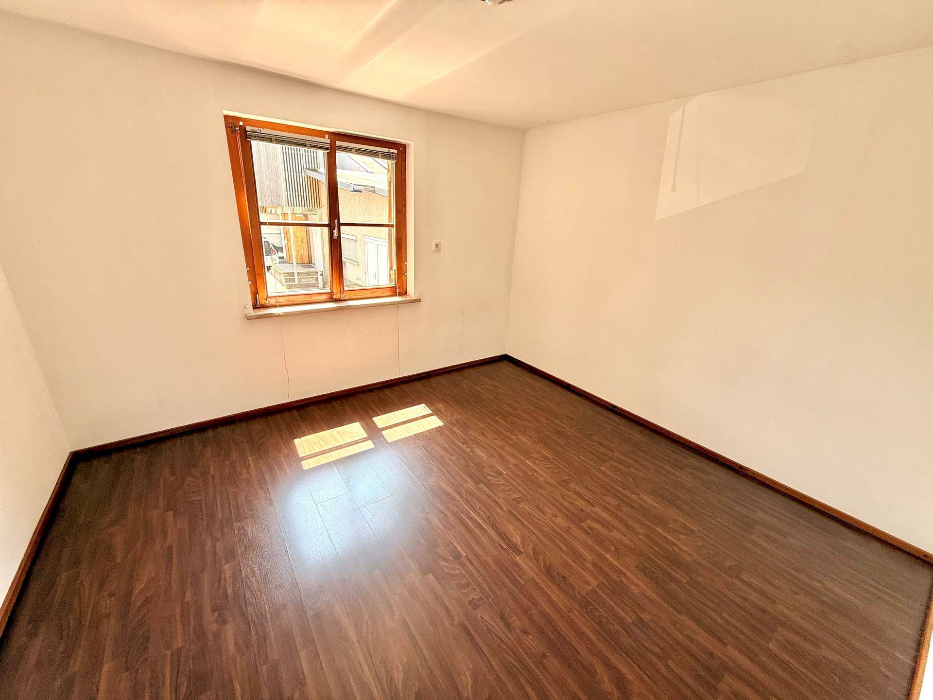 Leeres Zimmer mit dunklem Laminatboden und einem Fenster, das viel Tageslicht hereinlässt.