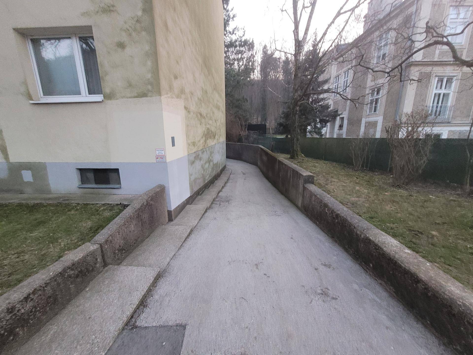 Betonierte Zufahrtsrampe entlang des Gebäudes mit Blick auf Bäume und weitere Häuser.