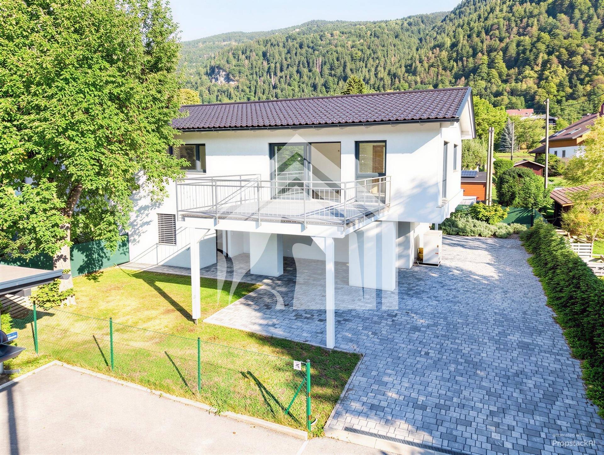 Modernes Einfamilienhaus mit weißer Fassade, großem Balkon und gepflasterter Auffahrt vor bergiger Kulisse.