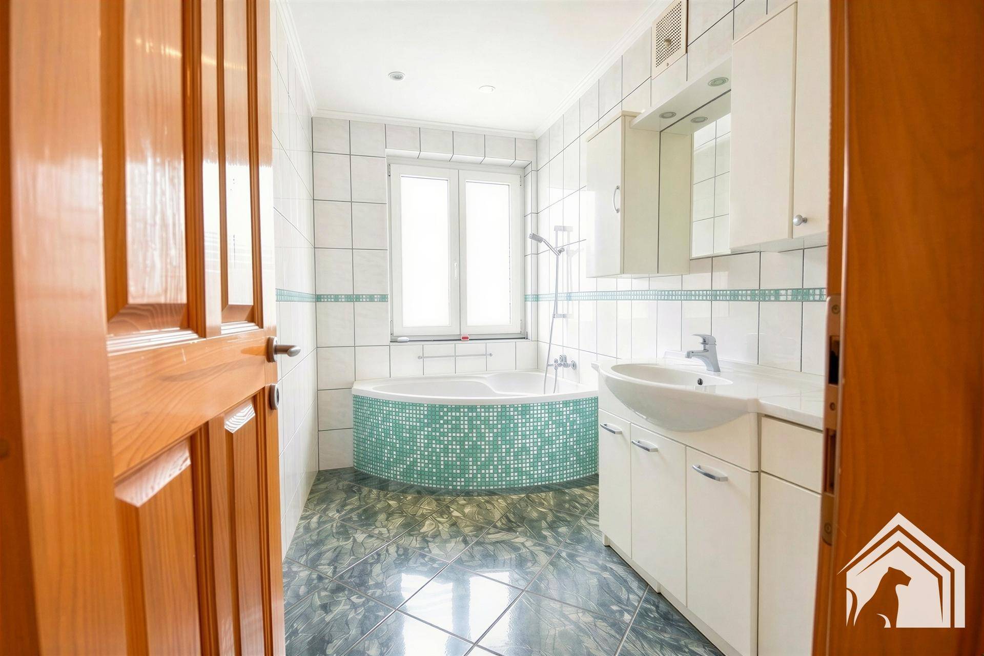 Badezimmer mit Eckbadewanne, Waschbecken und Fenster, hell gefliest in Weiß und Türkis.