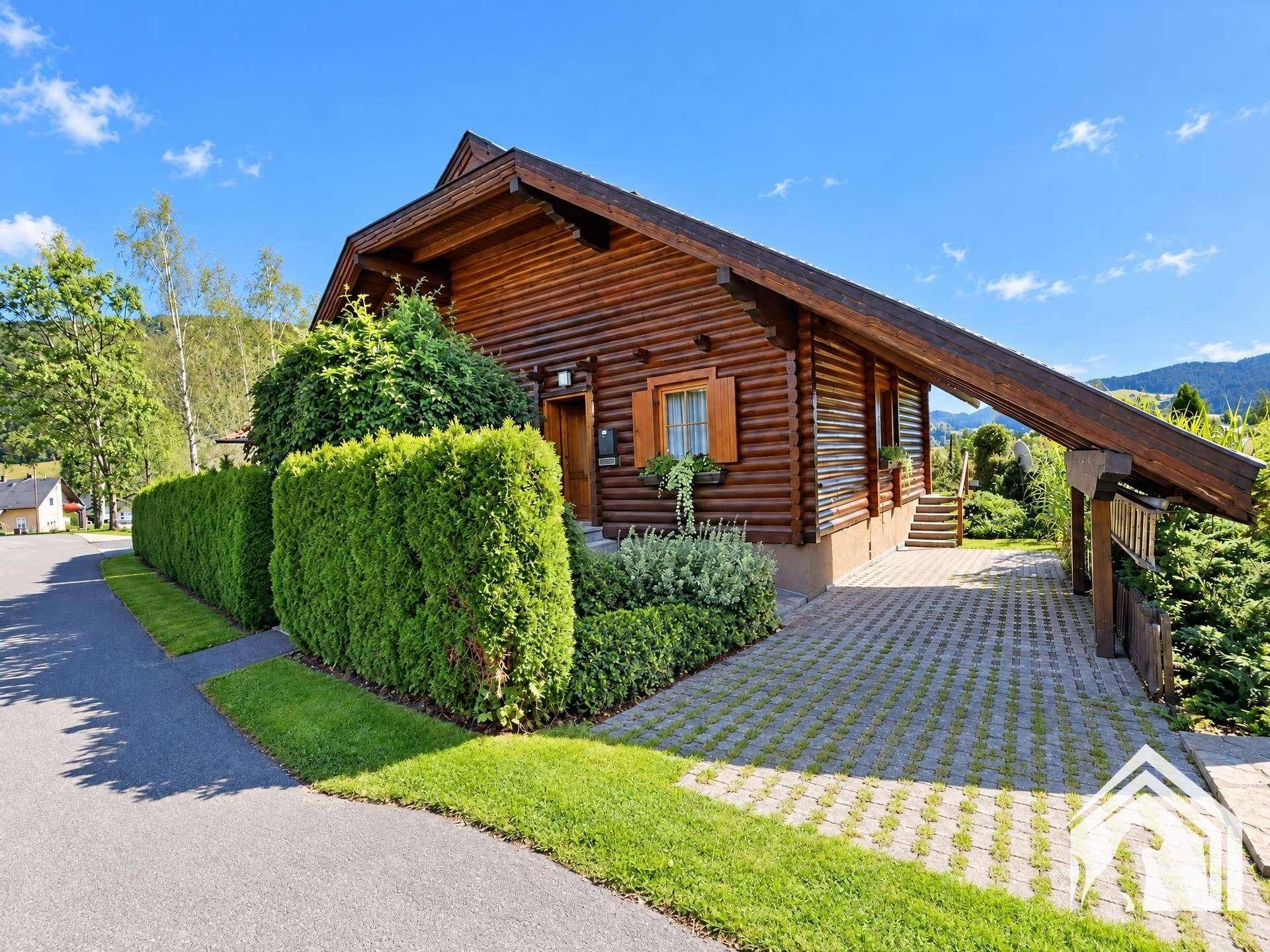 Seitenansicht des Holzhauses mit gepflasterter Auffahrt, umgeben von einer dichten Hecke und Bäumen.