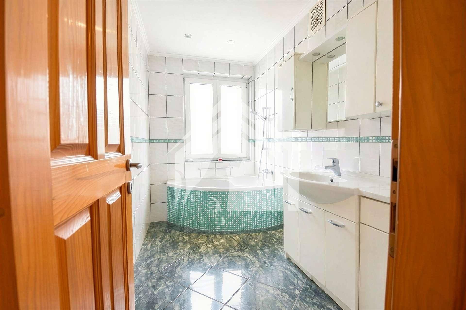 Badezimmer mit Eckbadewanne, weißen Fliesen und einem Fenster für Tageslicht.