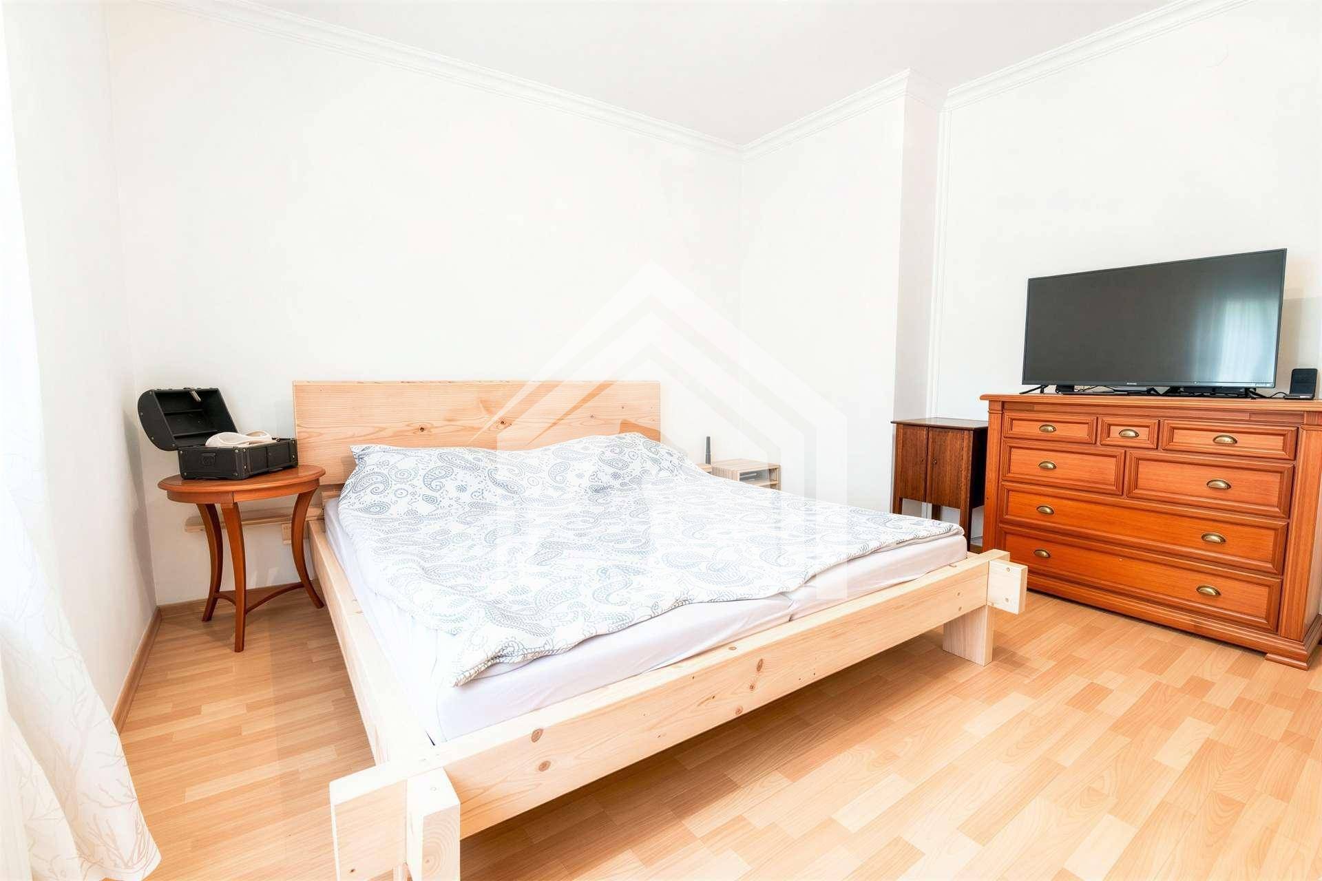 Schlafzimmer mit hellem Holzrahmenbett, Kommode und Fernseher.