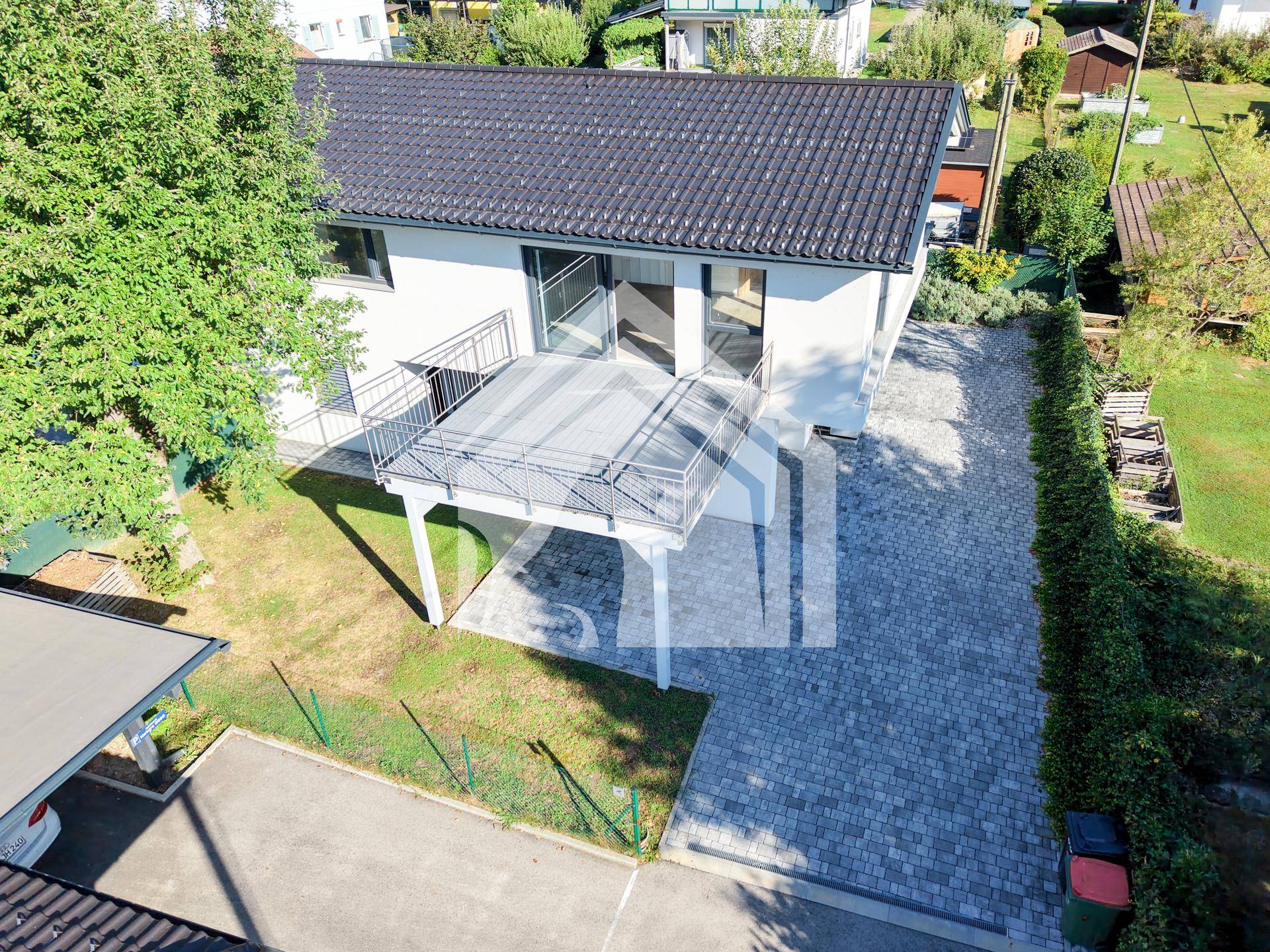 Vogelperspektive eines modernen Hauses mit großem Balkon, gepflasterter Auffahrt und umgebendem Grün.