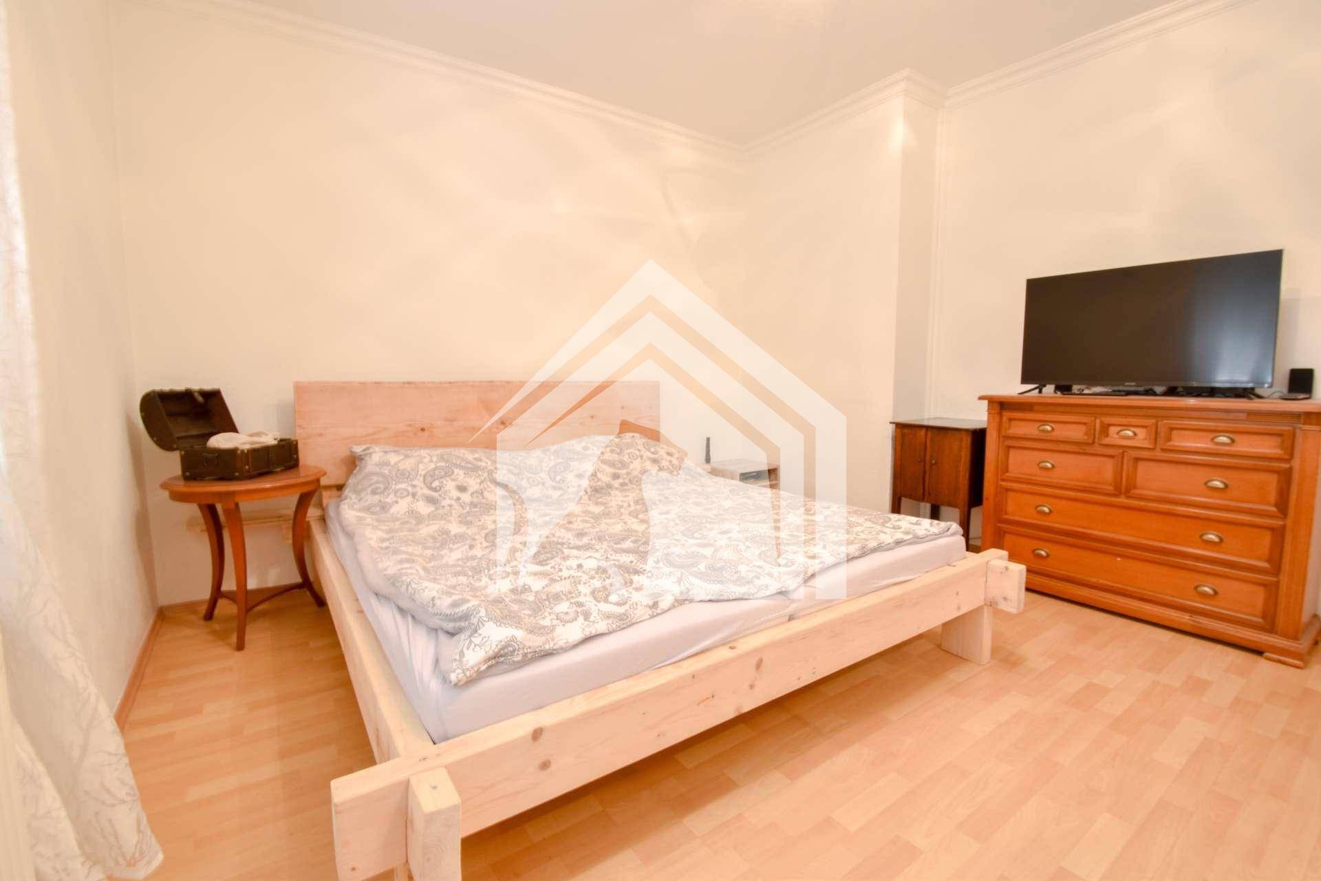 Schlafzimmer mit hellem Holzrahmenbett, Kommode und Fernseher.