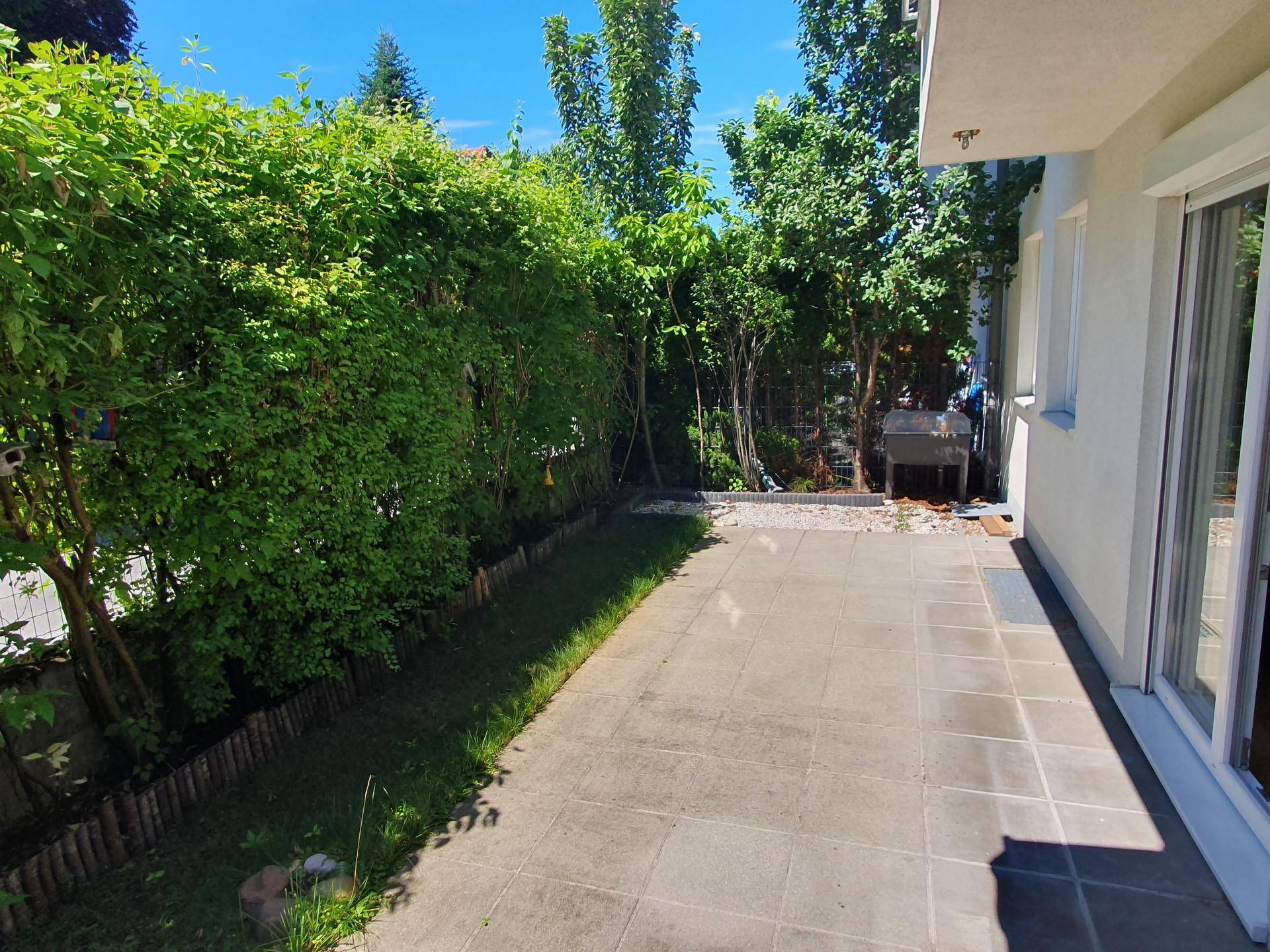 Gepflegter Gartenbereich mit Rasenfläche, Hecke und gepflastertem Sitzbereich mit Grill.