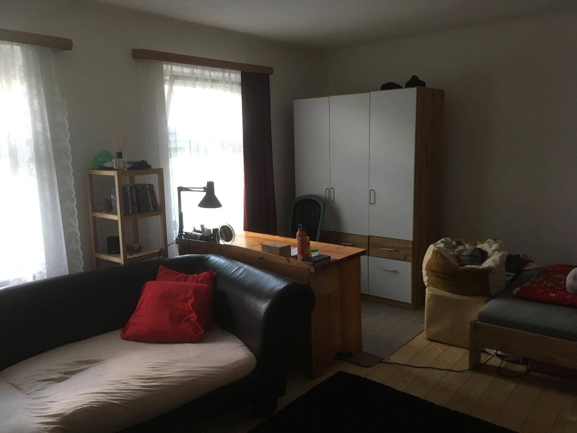 Wohnzimmer mit Sofa, Schreibtisch und Schrank, hell durch Fenster beleuchtet.