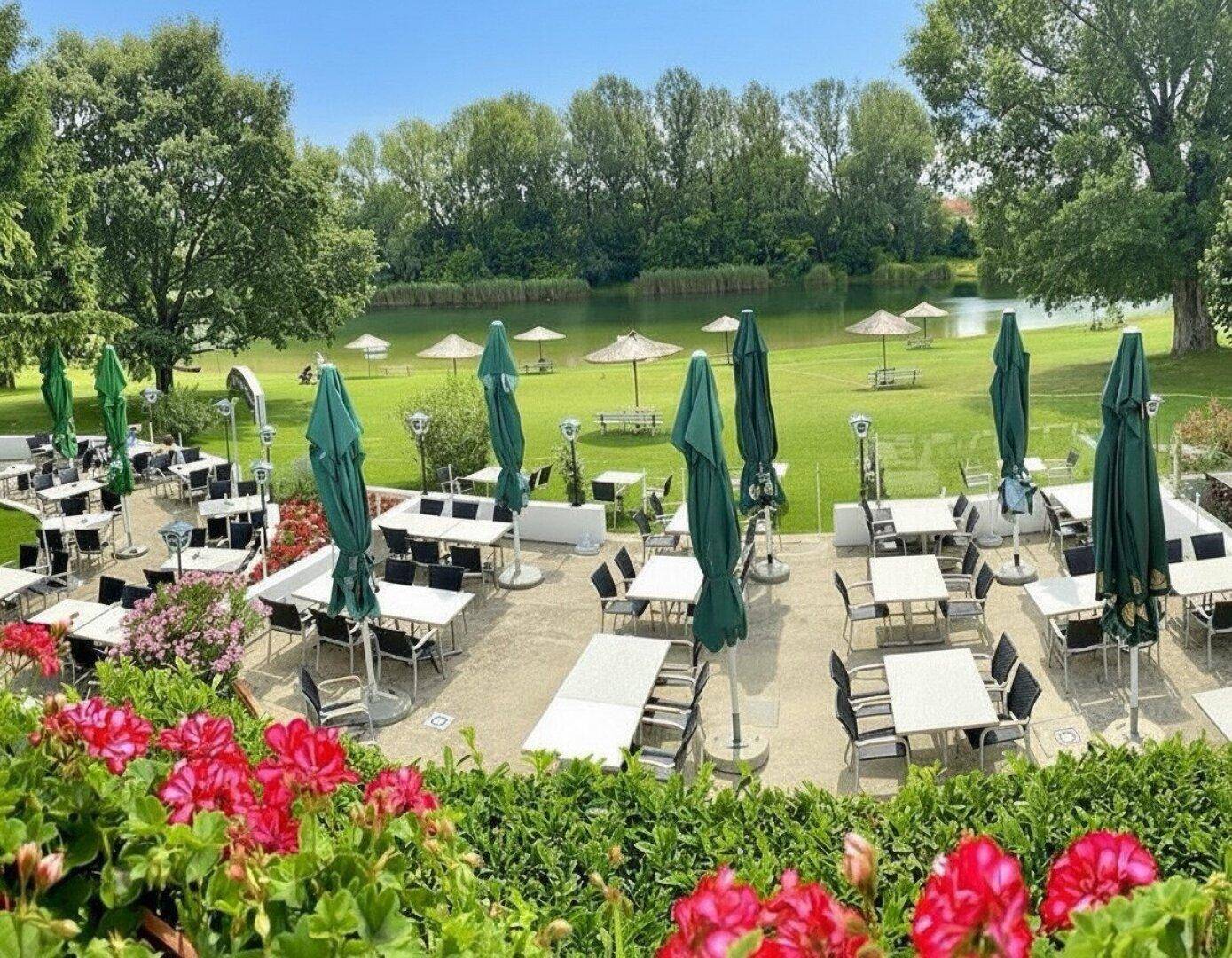 Große Außenterrasse eines Restaurants mit vielen Tischen und grünen Sonnenschirmen am Seeufer.