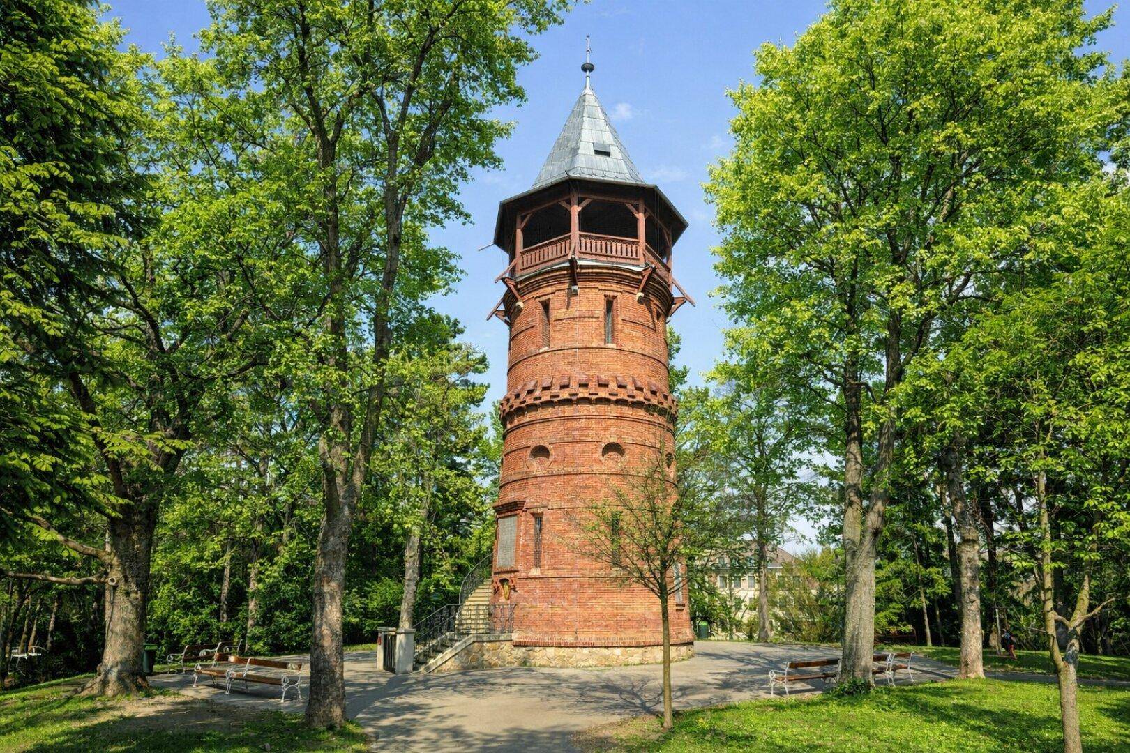 Historischer Backsteinturm mit Aussichtsplattform, umgeben von hohen Bäumen in einem Park.