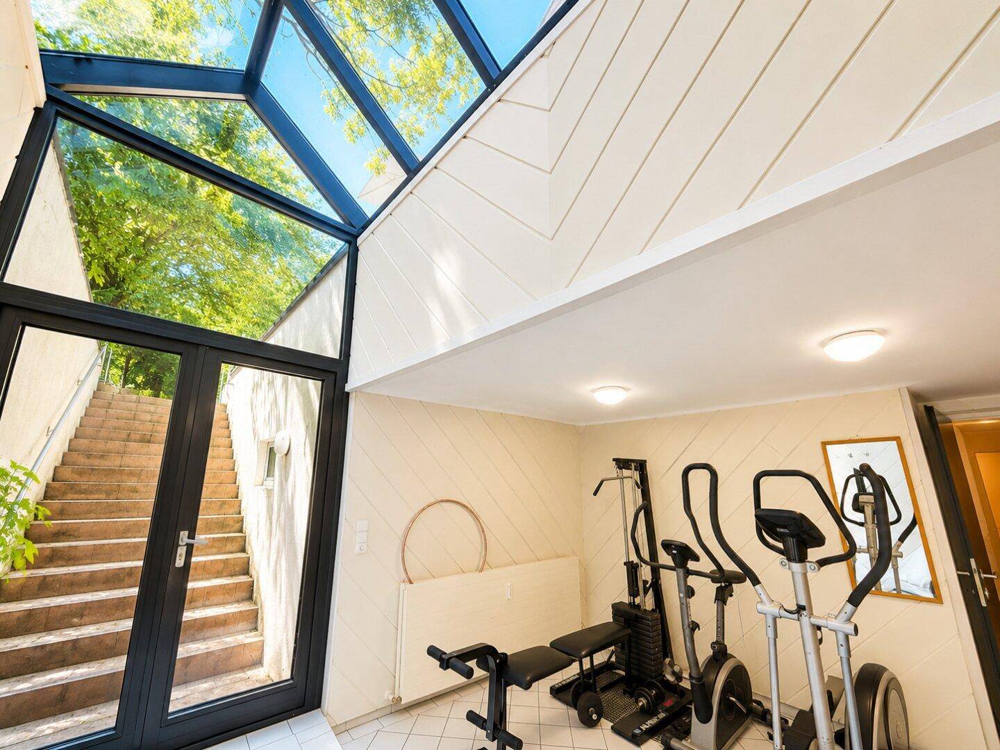 Fitnessraum mit Trainingsgeräten, großen Glasflächen und Zugang zu einer Außentreppe.