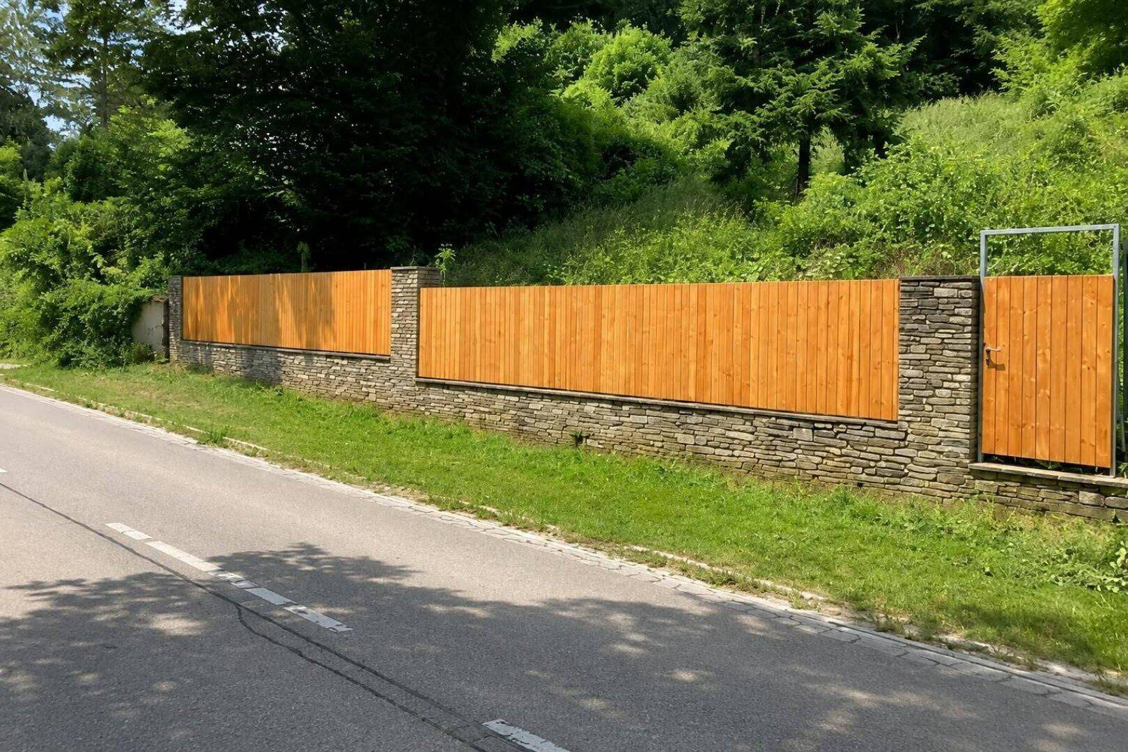 Langer Holzzaun mit Steinmauersockel und Tor entlang einer Straße, umgeben von Natur.