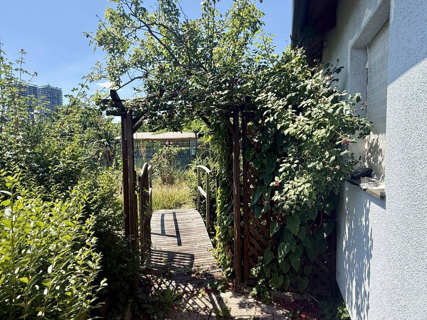 Idyllischer Gartenweg mit einem bewachsenen Holzbogen und kleinem Steg.