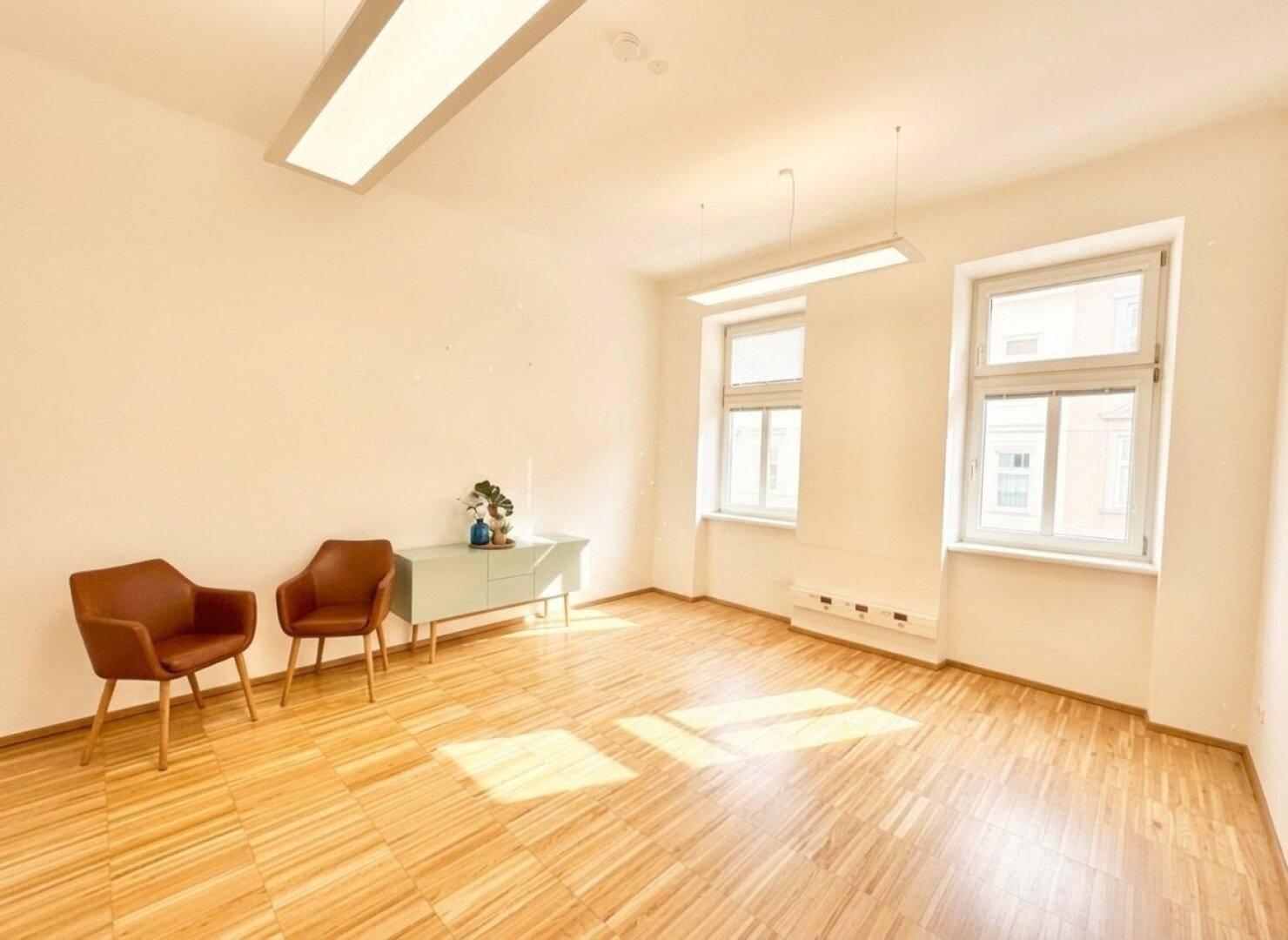 Heller Raum mit Holzfußboden und zwei Fenstern, ausgestattet mit modernen Büromöbeln und funktionaler Beleuchtung.