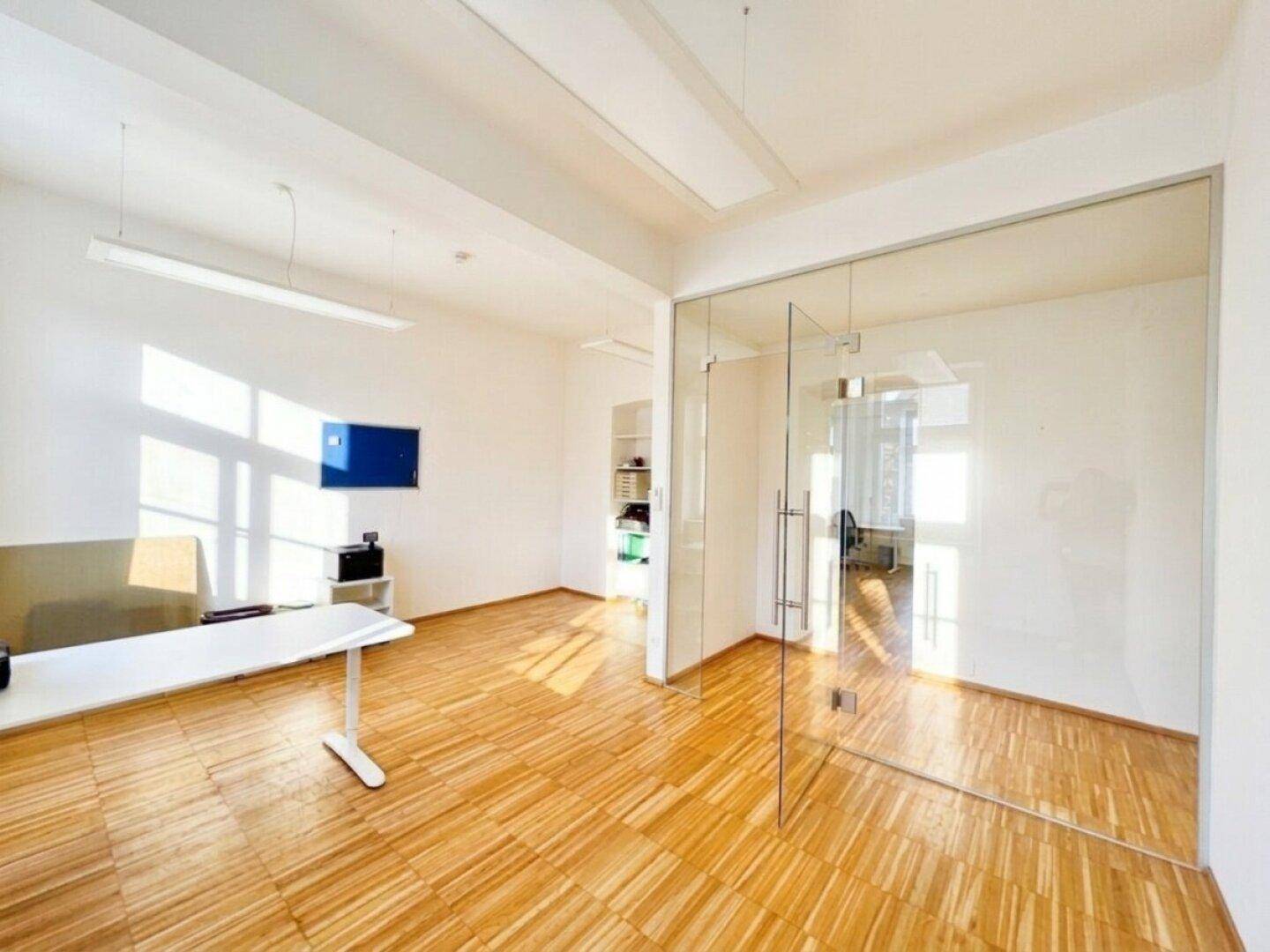 Modernes Büro mit einer eleganten Glaswand, hellem Holzfußboden und viel Tageslicht für eine offene Arbeitsatmosphäre.