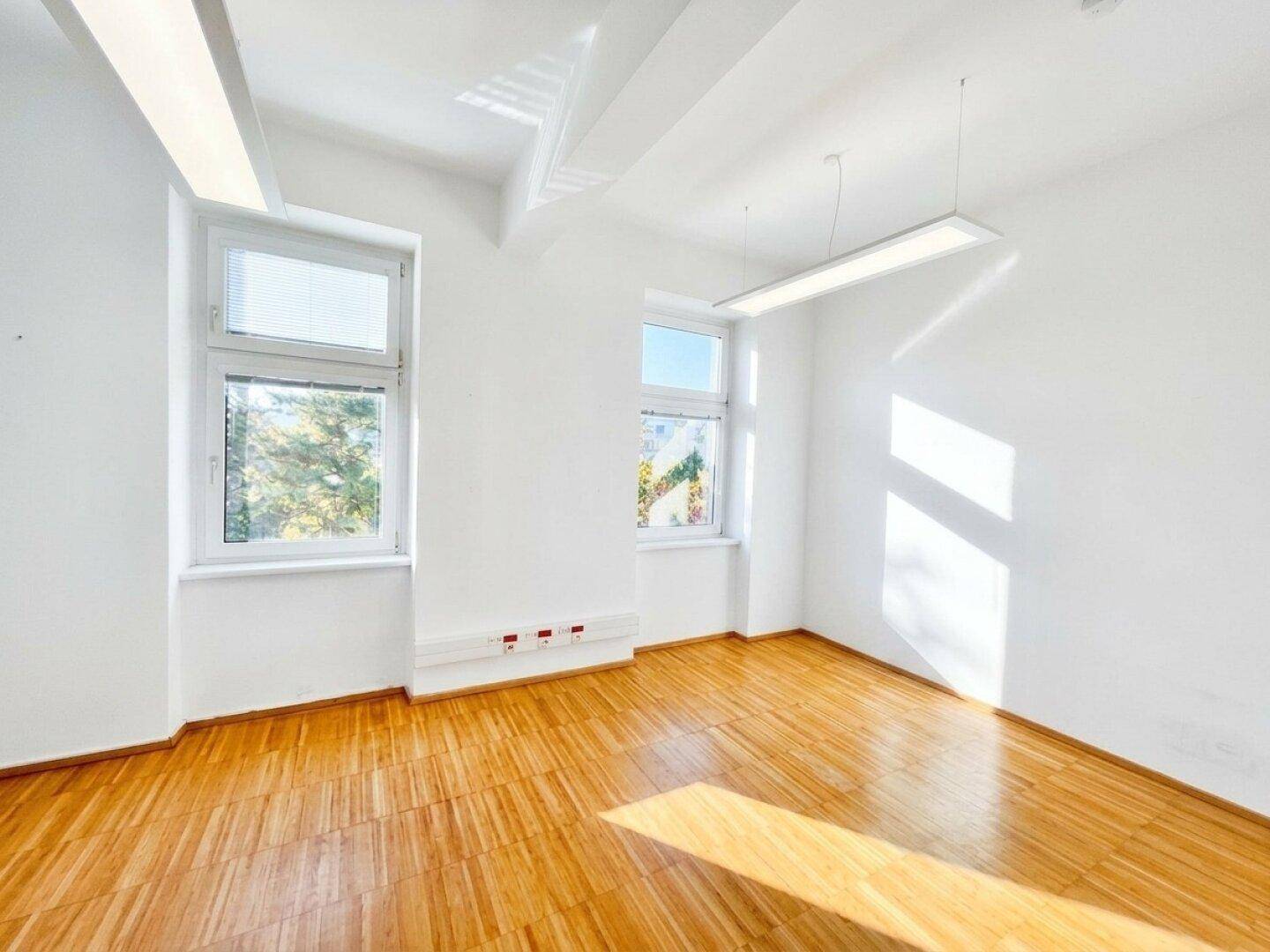 Leerer, heller Raum mit Holzfußboden und großen Fenstern, der viel Tageslicht und eine klare Atmosphäre bietet.