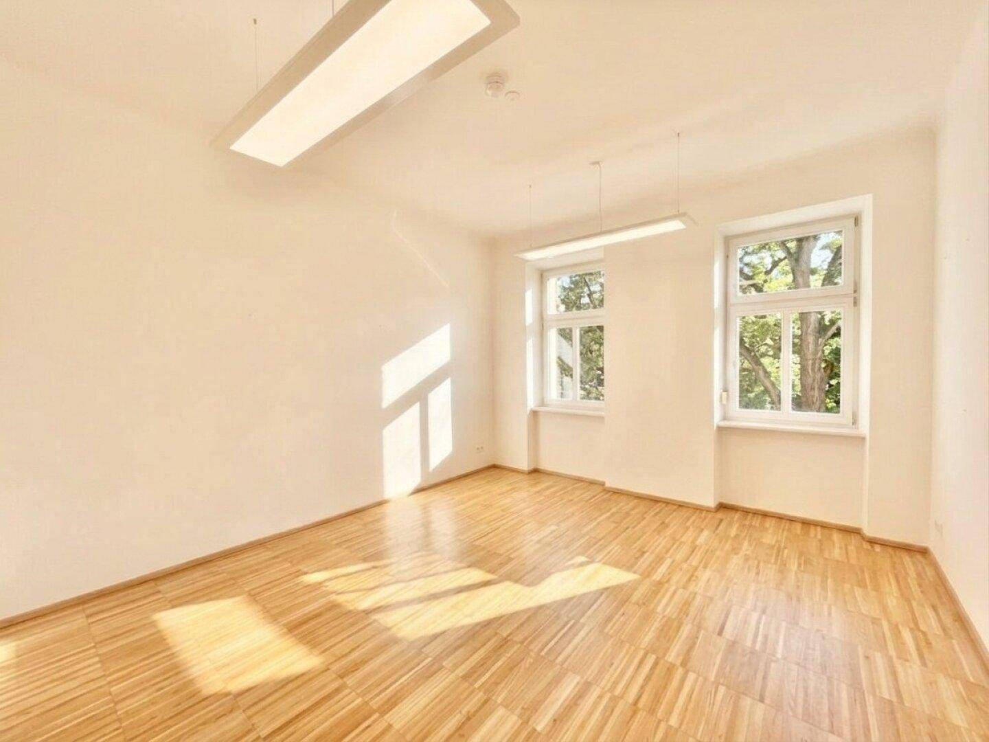 Geräumiger, heller Raum mit Holzfußboden und zwei großen Fenstern, ideal als Büro oder vielseitiger Wohnbereich.