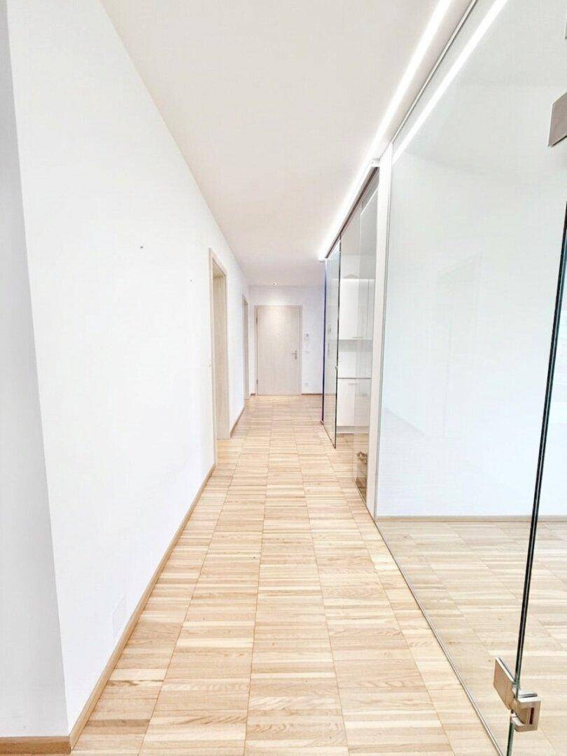 Langer, heller Flur mit Holzfußboden und einer modernen Glaswand, die den Blick in einen angrenzenden Raum freigibt.